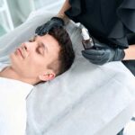 Nurse in protective gloves holding oil for skin and apply product on man face in salon